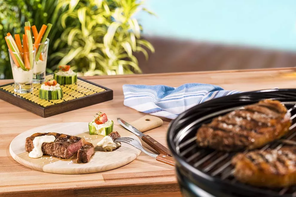 Duas peças de Contra filé em uma churrasqueira, no canto da imagem, com uma mesa de madeira ao centro, com uma tábua de cortar carne de madeira, com uma peça de Contra filé, com duas tiras cortados e um aperitivo de legumes ao lado, com alguns aperitivos de legumes ao fundo da mesa.