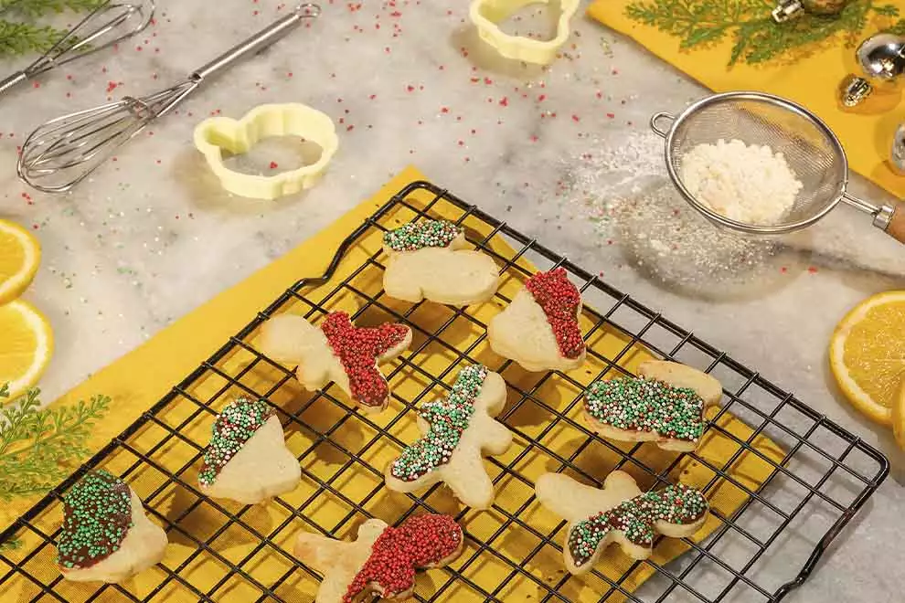 Diversos biscoitos natalinos em cima de uma forma retangular de grade, com diversos ingredientes ao redor, sob uma mesa de mármore.