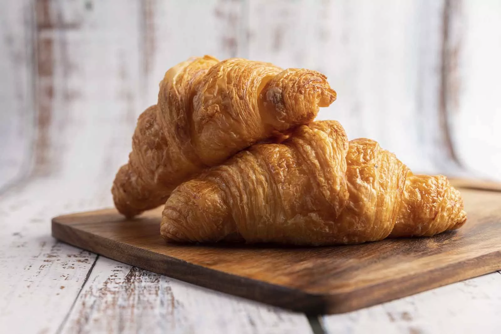 Imagem de duas unidades de Croissant um em cima do outro, com ambos sob uma tábua de madeira retangular, em cima de uma superfície de madeira 