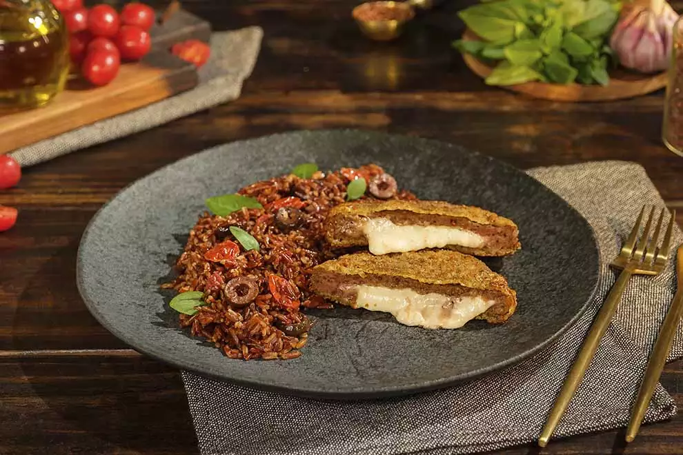 Imagem de um prato escuro com arroz temperado e um polpetone cortado ao meio, com um par de talheres ao lado, em cima de uma mesa de madeira. 