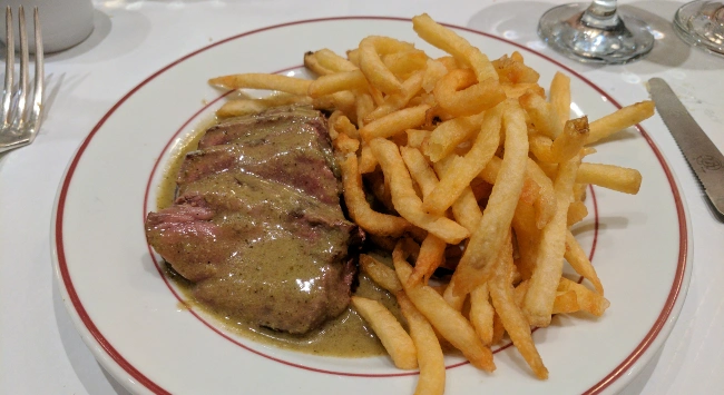 Bife fatiado, servido com batatas fritas e molho em um prato branco com bordas vermelhas.