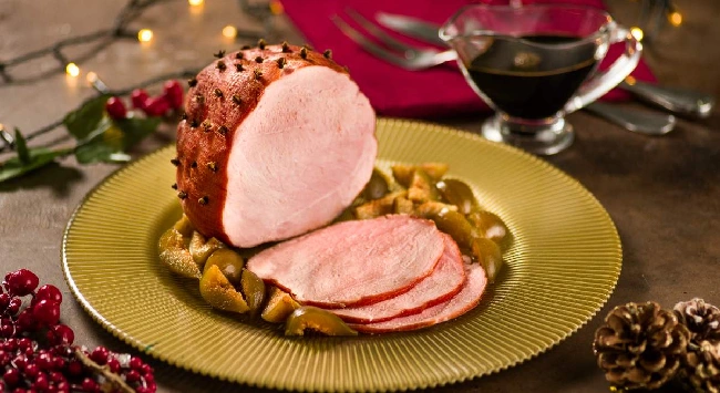 Tender bolinha fatiado, servido com legumes em um prato dourado redondo e bem raso. O fundo da mesa está com decorações de Natal e piscas piscas.