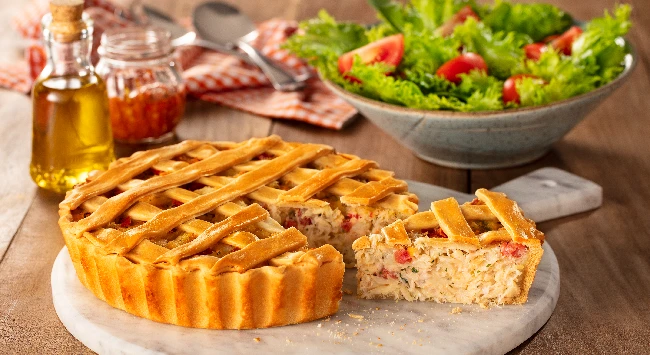 Torta de bacalhau cremosa em uma tábua de cerâmica branca. Ao fundo, uma salada de folhas verdes com tomates e um vidro de azeite.