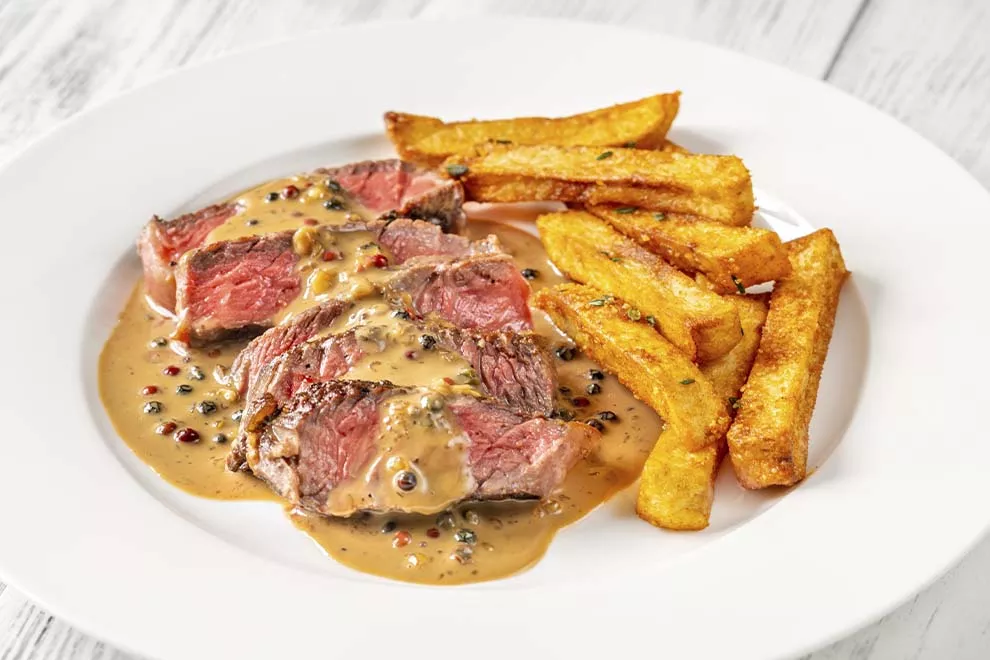 Filet au poivre, cortado em tiras, com batatas fritas ao lado, sob um prato redondo branco, em cima de uma mesa de madeira.