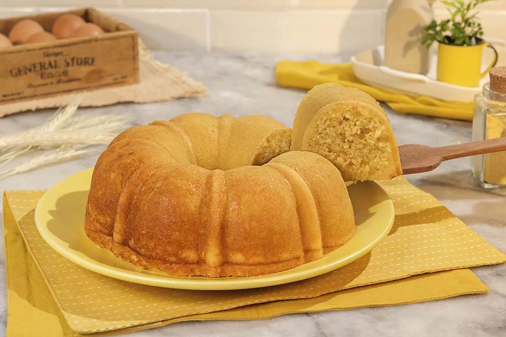 Imagem de um bolo de fuba em cima de prato redondo amarelo, com uma fatia dele sendo retirada, sob uma mesa de mármore