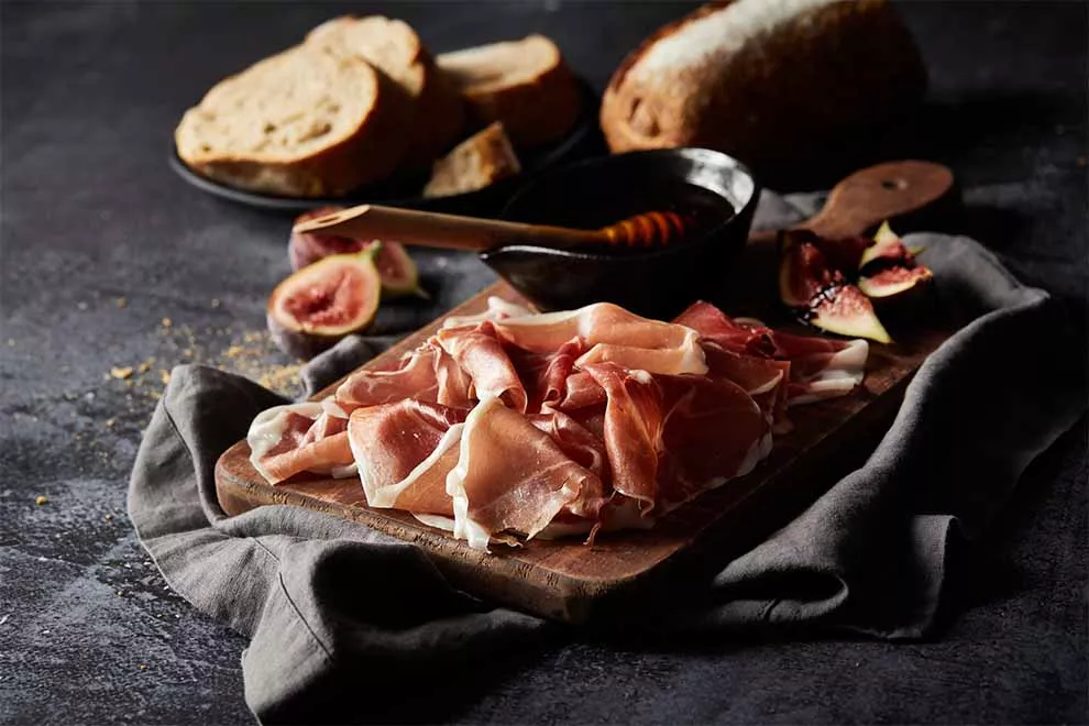 presunto parma em cima de uma tábua de madeira com um pequeno bowl com mel e alguns pedaços de figo ao lado