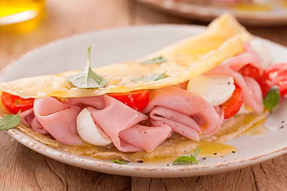 crepioca com presunto, muçarela de bufula e manjerona dentro de um pequeno prato de porcelana branco em cima de uma mesa de madeira