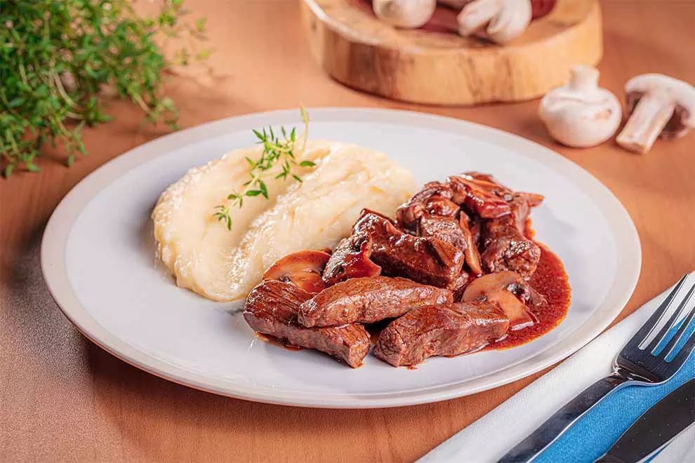 filé mignon ao molho madeira com cogumelos e purê de batata dentro de um prato branco de porcelana com talheres ao lado em cima de um pano branco e azul em cima de uma mesa