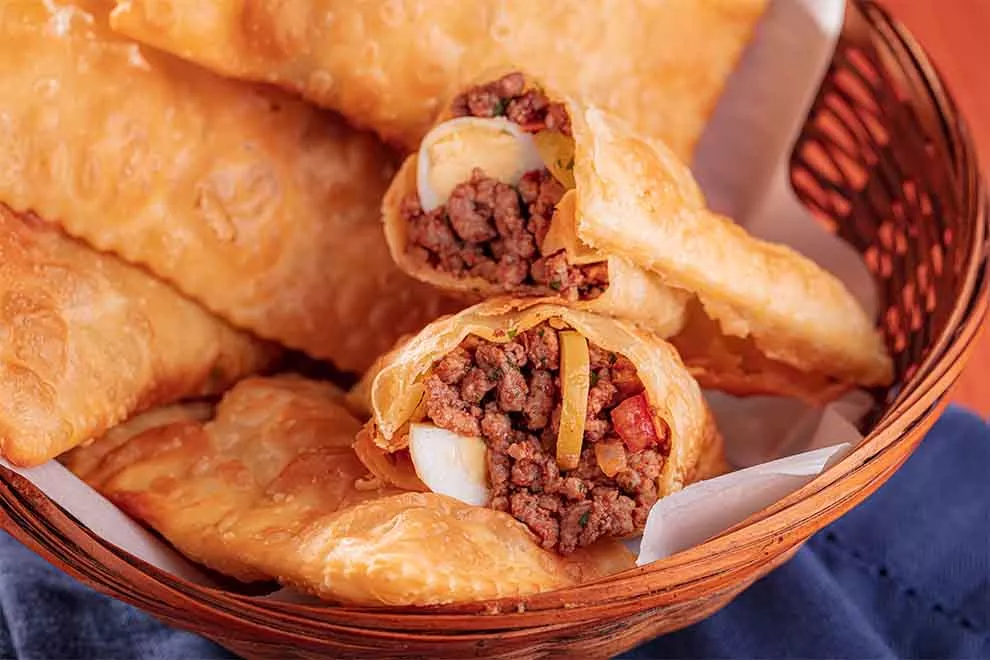 vários pastéis pequeno de carne moída dentro de uma pequena cesta com guardanapos com um pano azul escuro em baixo da cesta em cima de uma mesa