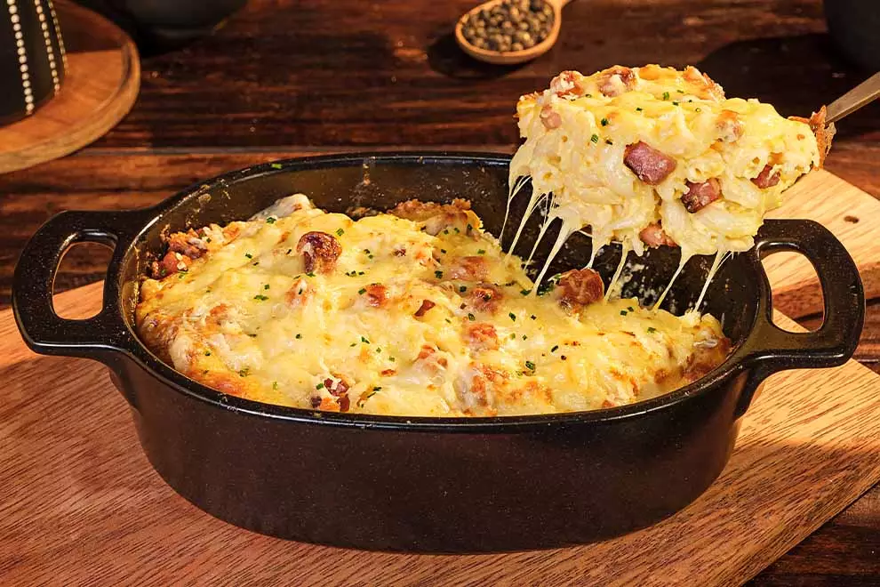 Macarrão de forno com bacon e queijo, sob uma panela escura, com uma colher retirando uma parte do Macarrão, sob uma mes de madeira.