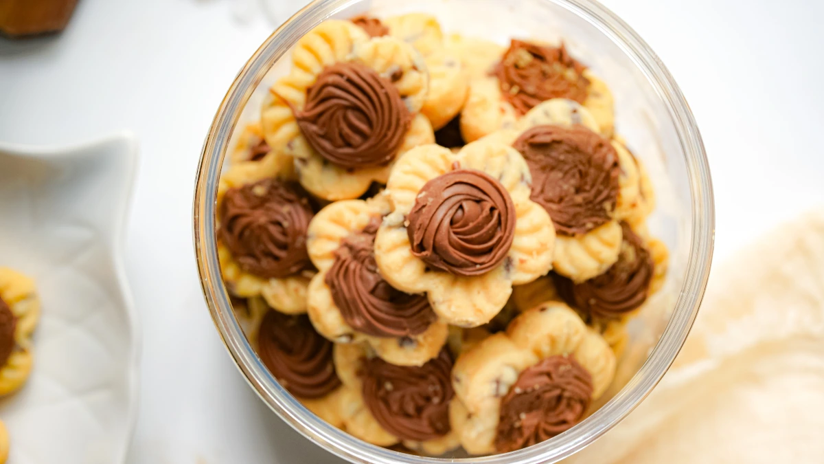 Biscoitos amanteigados em formato de flor com detalhes de chocolate decorado no centro, armazenados em um pote transparente.