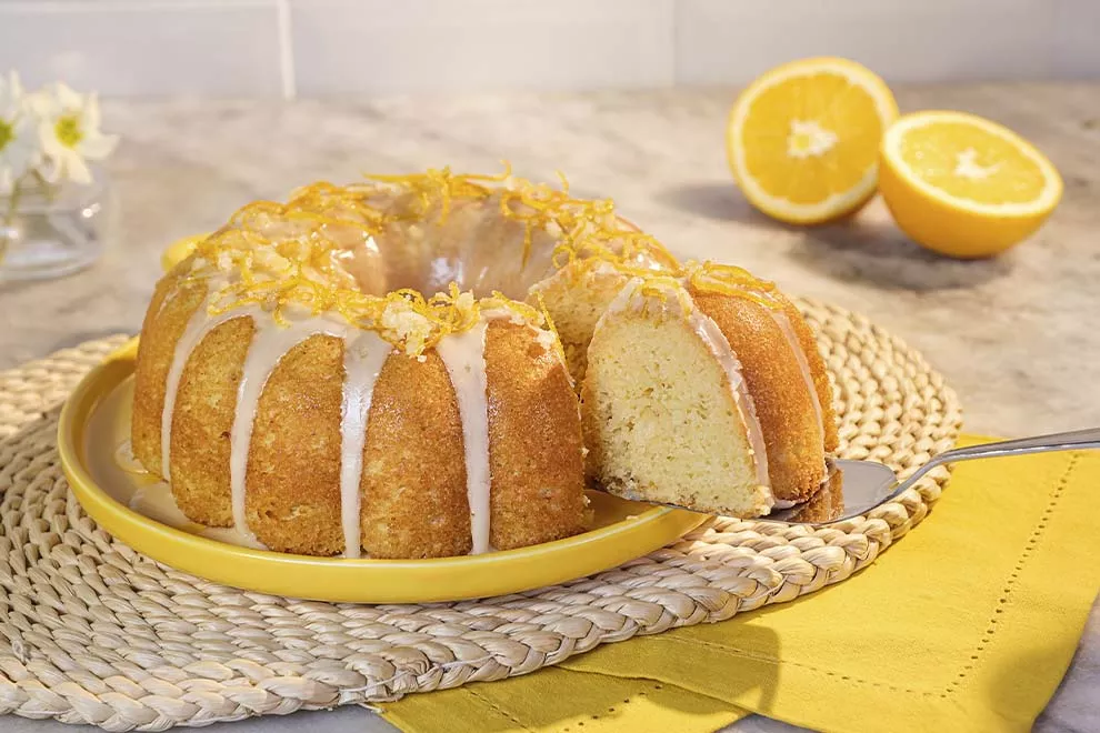 Imagem de um bolo de laranja com cobertura em cima de um prato redondo amarelo, com uma fatia dele sendo tirada, sob uma mesa de mármore