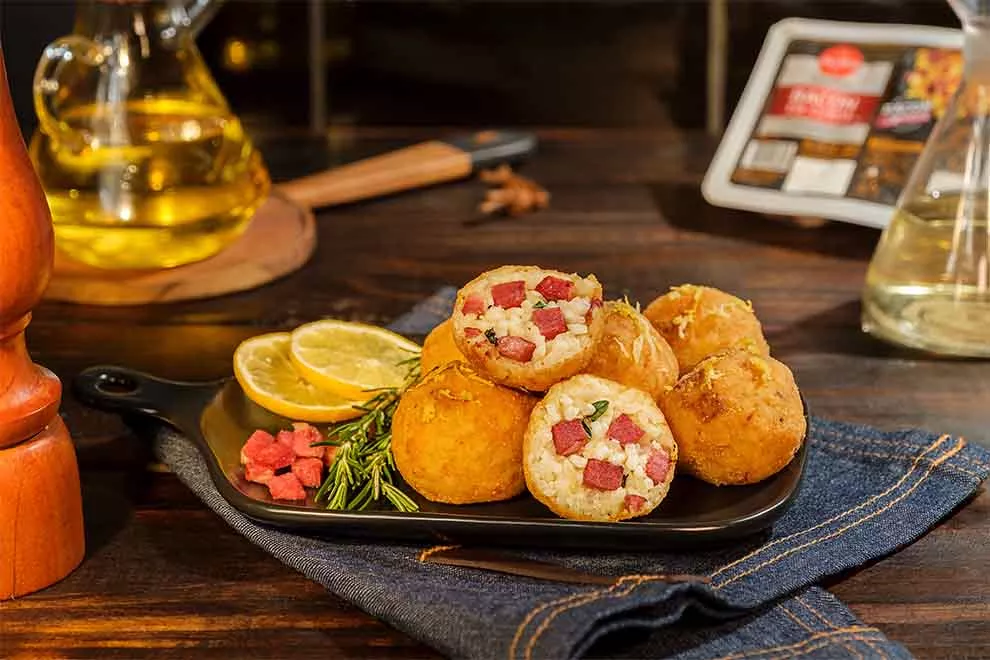 arancini de bacon com dois ramos de alecrim e duas rodelas de limão dentro de um pequeno prato retangular de porcelana preto em cima de um guardanapo jeans em cima de uma mesa