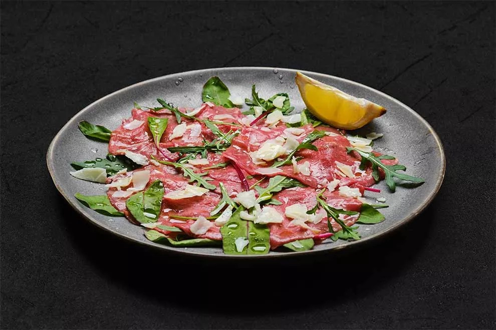 carpaccio dentro de um prato redondo de porcelana cinza, com um pedaço de limão e algumas folhas em cima de uma superfície preta e fundo preto 