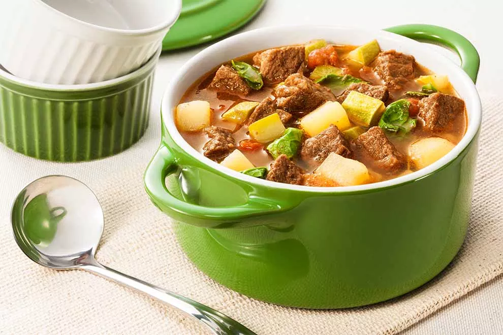 sopa de coxão duro com legumes dentro de uma panela funda verde, pequeno bowls ao fundo e uma concha ao lado, em cima de um pano bege na mesa branca