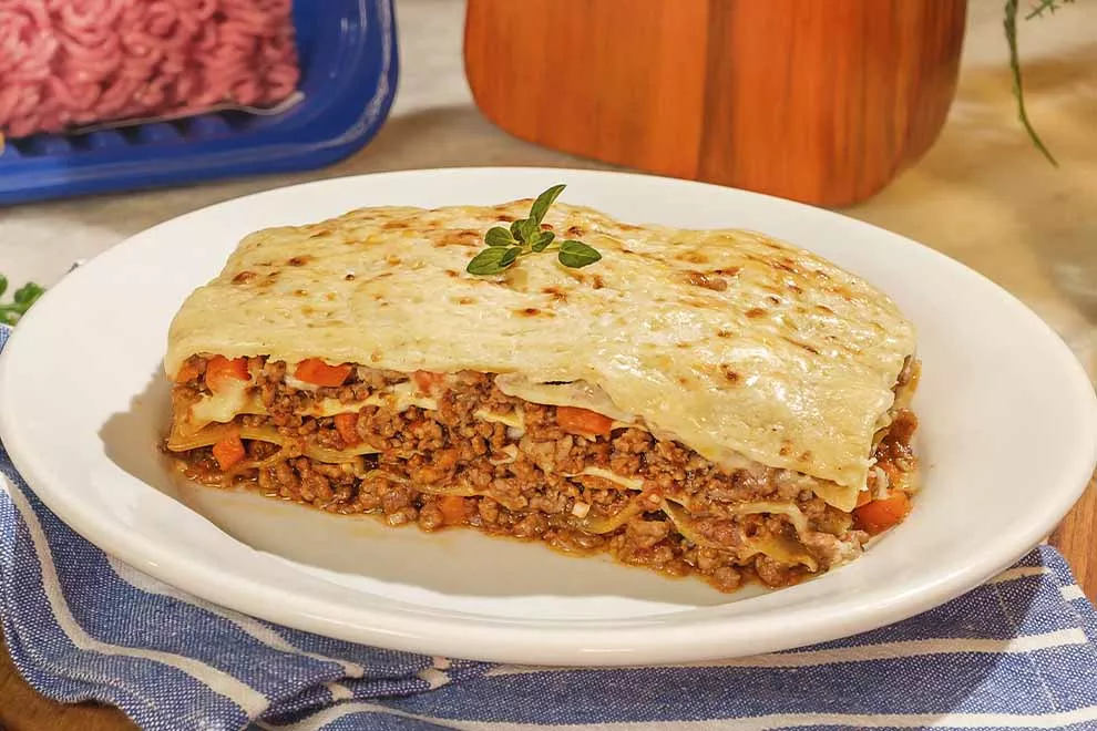 Lasanha de carne moída em cima de um prato redondo branco, sob uma mesa de mármore.