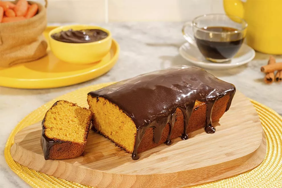 Bolo de cenoura com chocolate vegano, com uma fatia dele cortado, em cima de uma tábua de madeira, sob uma mesa.