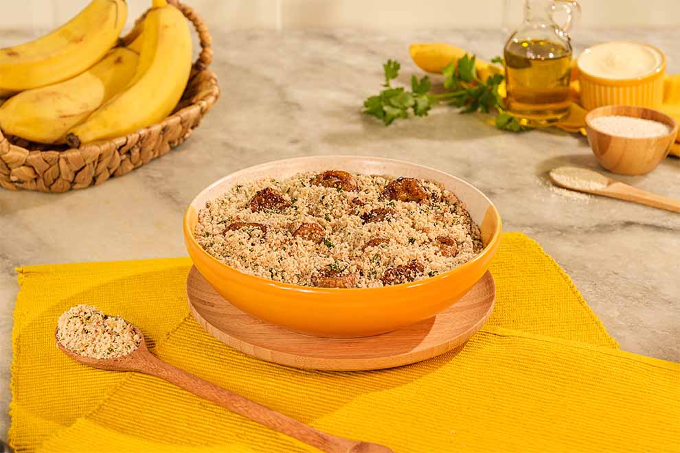 um pequeno bowl de porcelana com farofa de banana dentro e com um pequeno prato fundo de bambu em baixo, ao lado com uma colher de madeira e farofa dentro em cima de um pano amarelo e em cima de uma mesa 