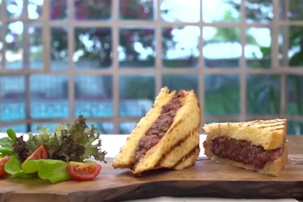 sanduíche de tartare de patinho em cima de uma tábua de madeira e em cima de uma mesa branca