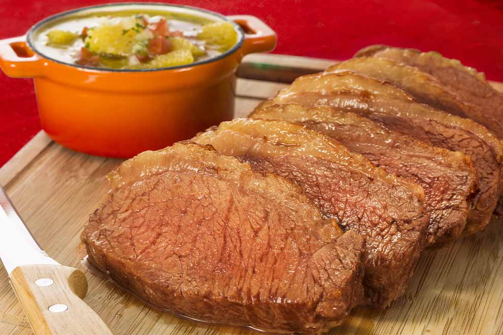 Imagem de diversas fatias de picanha assada, sob uma tábua de madeira, com uma faca e uma combuca com mandioca cozida ao lado, em cima de uma mesa com pano vermelho. 