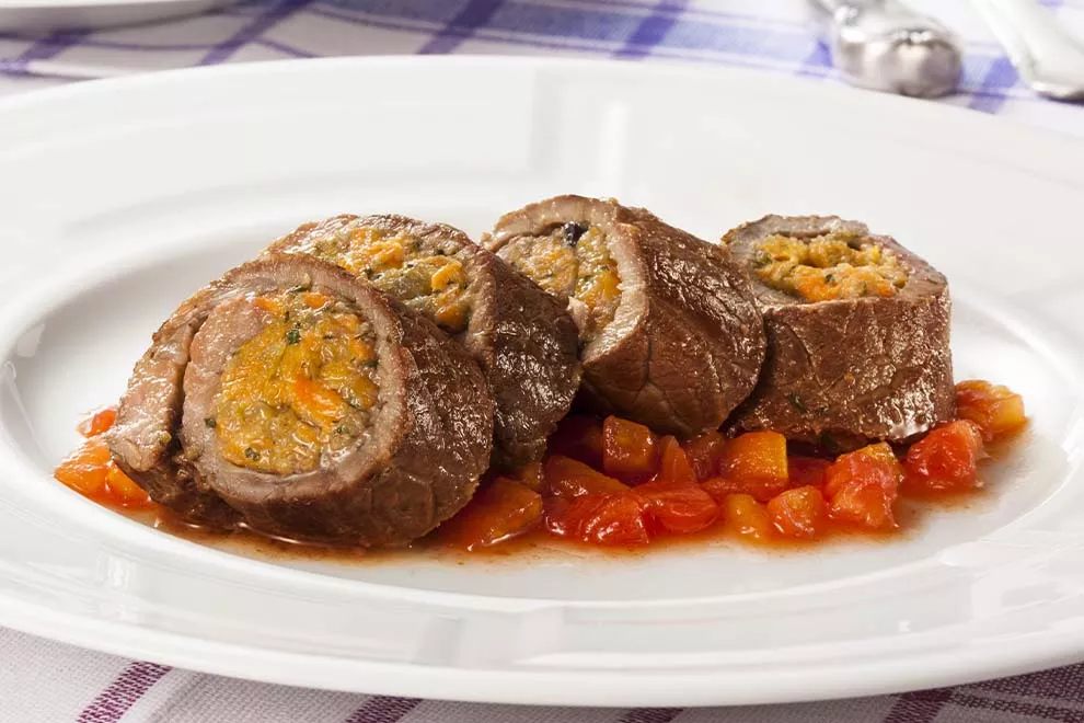 Quatro unidades de Bife à rolê, em cima de um prato redondo branco, sob uma mesa com toalha quadriculada.