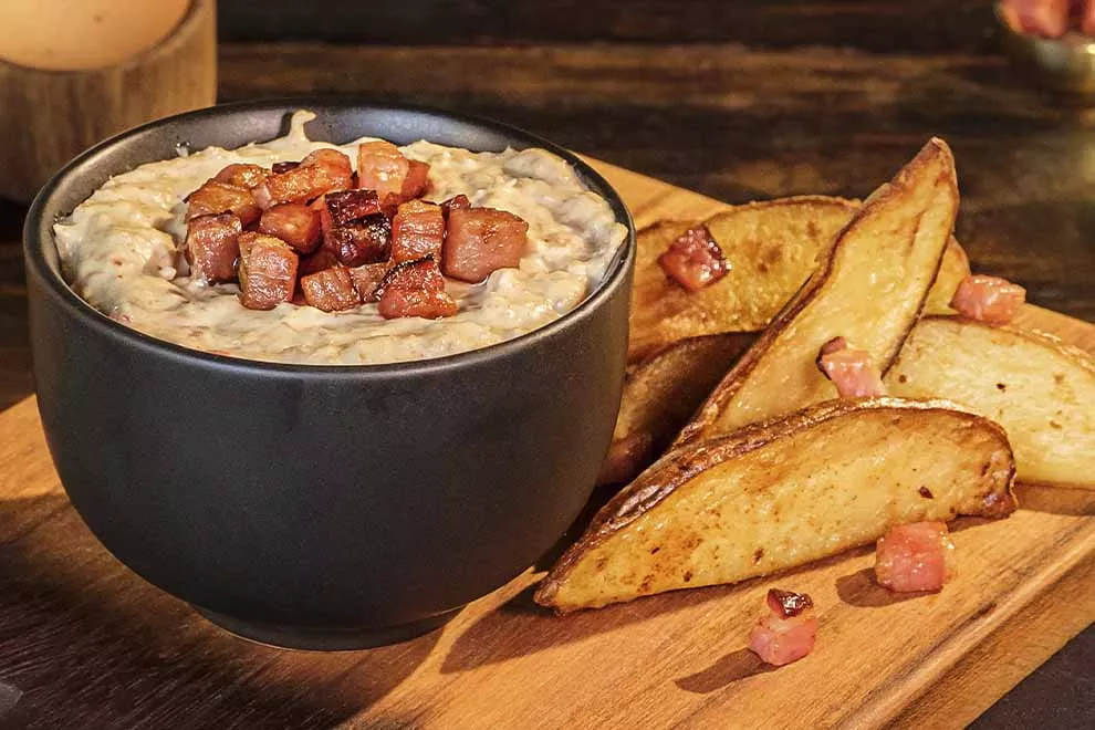 Maionese de bacon em cima de uma tigela escura, com algumas batatas rústicas ao lado em cima de uma tábua de madeira, sob uma tábua de madeira. 