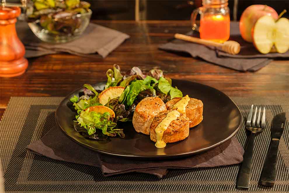 filé mignon suino com mostarda e mel, com uma salada ao lado dentro de um prato preto de porcelana em cima de um pano escuro e de um jogo americano retangular escuro com talheres ao lado na mesa