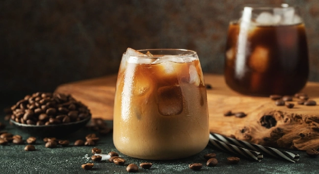 Copo com café gelado proteico servido em uma mesa escura com grãos de café ao lado.