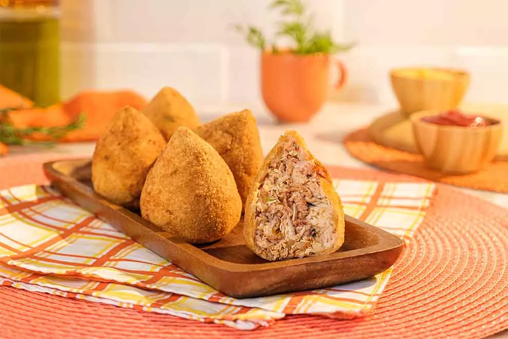 coxinha sem massa de frango dentro de um pequeno recipiente de madeira em cima de um pano xadrez em branco, amarelo e vermelho em cima de um jogo americano redondo alaranjado na mesa