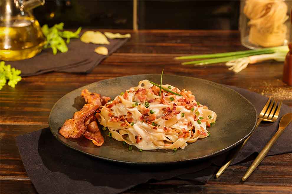 macarrão ao molho branco e bacon com cebolinha em cima dentro de um prato preto de porcelana com talheres ao lado em cima de um pano escuro e em cima de uma mesa