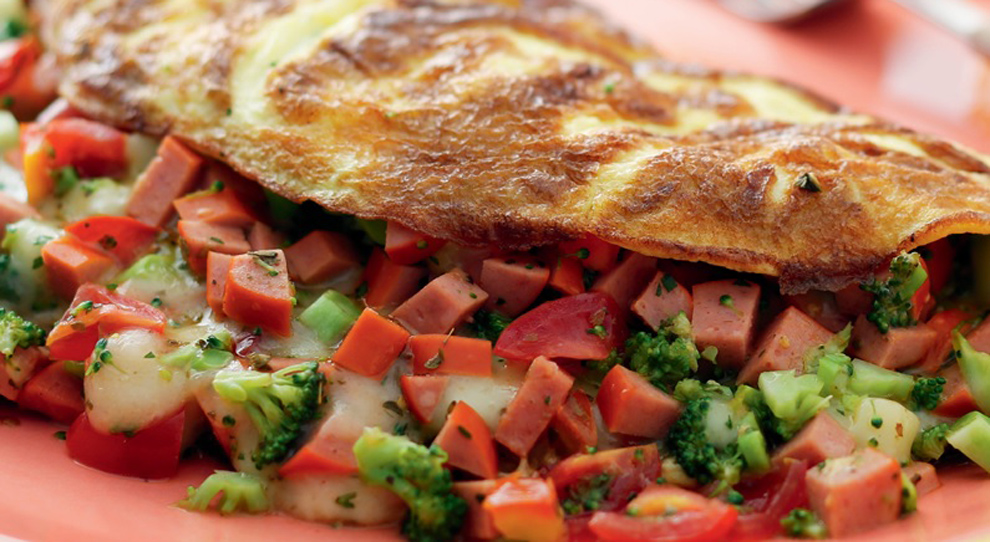 Receita de omelete com ingredientes variados. Uma omelete dourada e macia, dobrada ao meio, recheada com salsicha picada, brócolis, tomate, cenoura e queijo derretido. O prato colorido e apetitoso está servido sobre um prato laranja, destacando os ingredientes frescos.