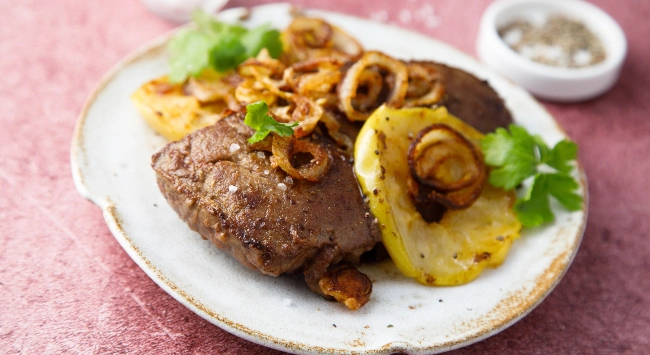 Prato de fígado acebolado servido com rodelas de batata douradas e cebolas fritas, decorado com salsa.