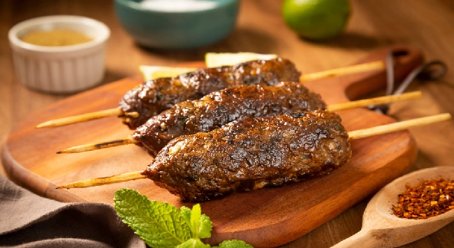 Espetinhos de kafta de carne em uma tábua de madeira com molho de iogurte e mostarda ao fundo.